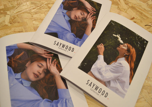 Saywood lookbooks in recycled paper on recycled wood table. Books have two different covers, one model in the blue Edi shirt, recycled cotton, with her hands above her head. 2nd model wears white shirt Edi holding a flower