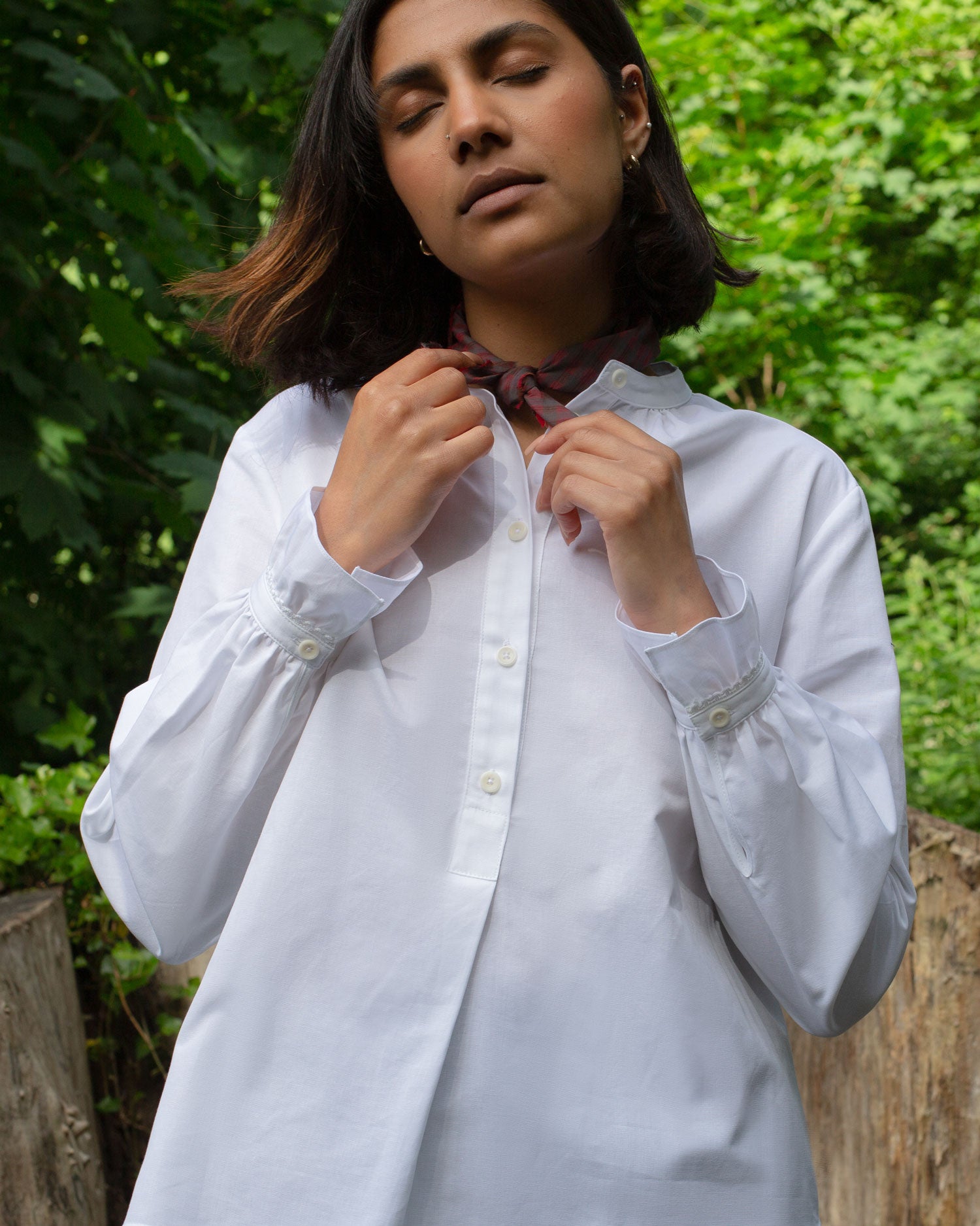 Model has eyes closed and is wearing Saywood's white recycled cotton blouse, Marie Blouse. She pulls at a red check neck tie at her neck, and the frilled cuffs are visible.
