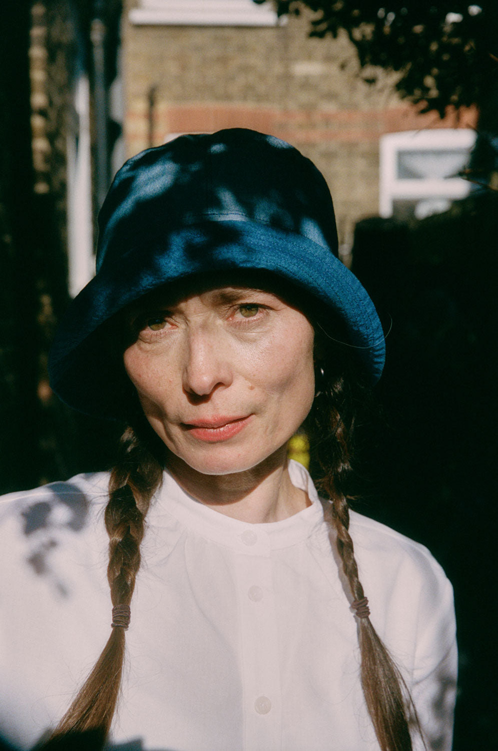 Close up of a woman standing in dappled light, with plaited pigtails. She wears Saywood's Japanese denim Reversible Bucket Hat and white recycled cotton gathered neck Marie Blouse.