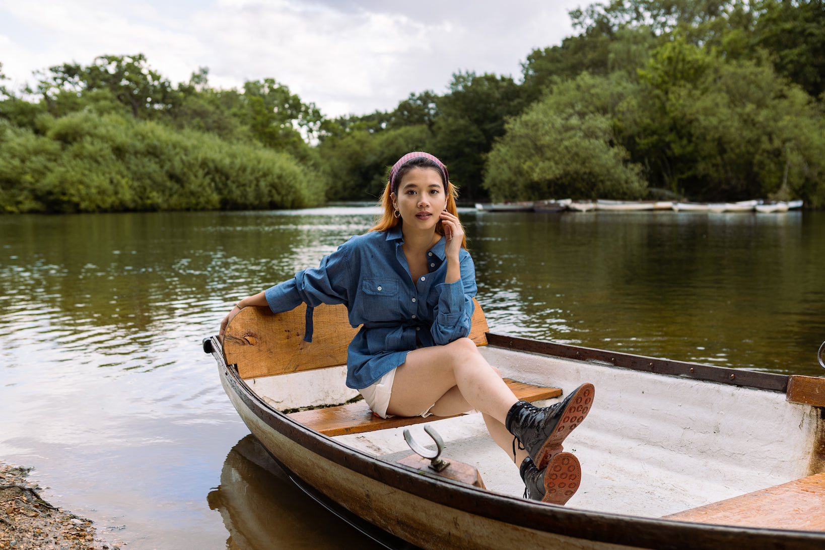 Model sits in row boat on lake, with foot stacked on top of other foot. She wears Saywood's Zadie Boyfriend Shirt in Japanese Denim, white shorts and black boots with a red sole.