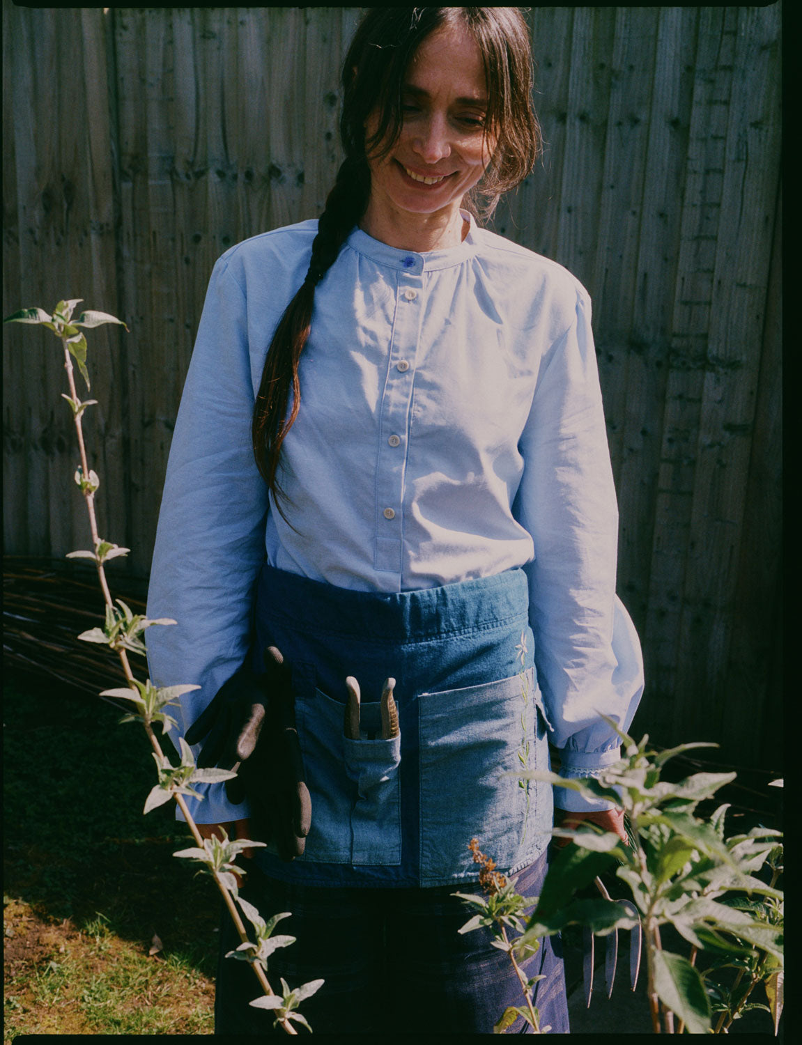 A woman stands smiling in her garden, looking at plants. Around her waist is the Multifunctional Gardening Tool Belt Apron in Japanese Denim with gardening hand tools in. She is wearing Saywood's Marie A-line Blouse with gathered neck and cuffs, with a blue vintage glass button at the neckline.