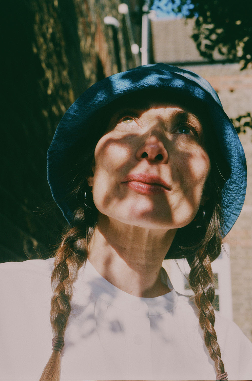 Close up of a woman standing in dappled light, looking up, with plaited pigtails. She wears Saywood's Japanese denim Reversible Bucket Hat and white recycled cotton gathered neck Marie Blouse.