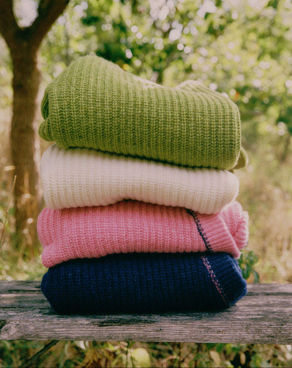 Stack of colorful sweaters in a natural setting with trees and greenery.