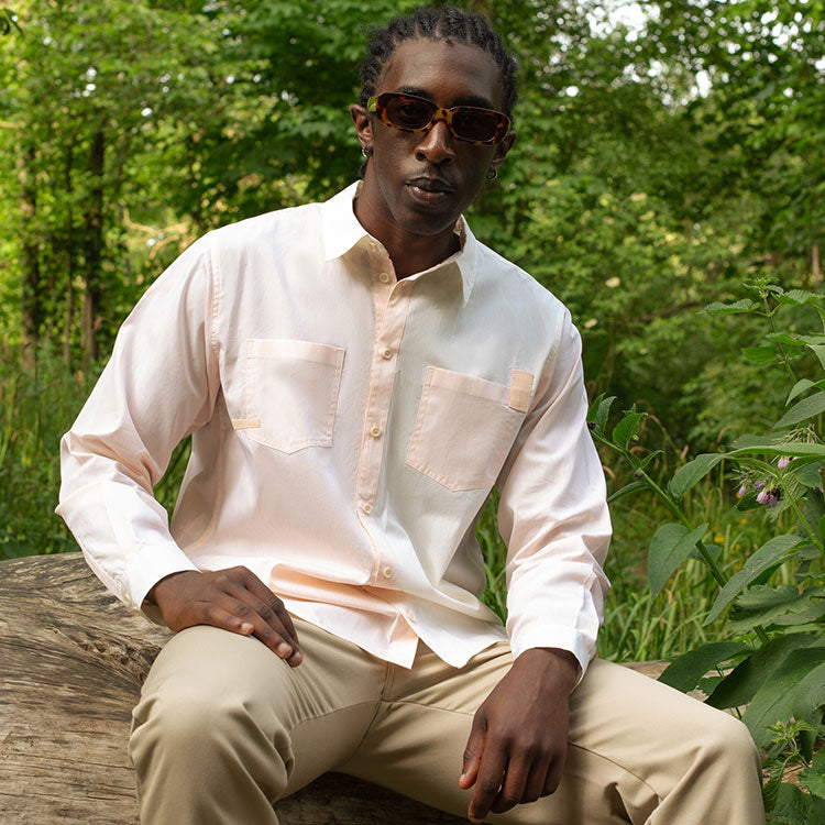 Model sits on a log surrounded by nature. He wears Saywood's Eddy Men's Colourblocked Shirt in soft orange and lemon, with tan trousers and sunglasses.