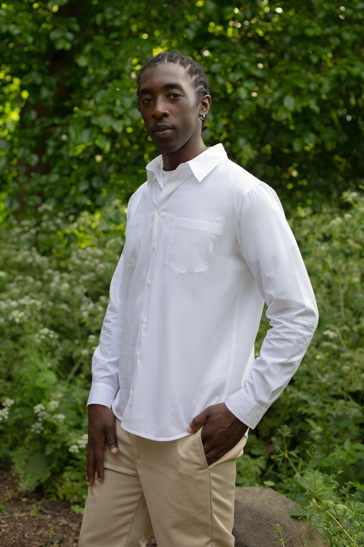 Model wears Saywood's men's white patch pocket shirt in Supima cotton and bamboo. He stands with one hand in his pocket, surrounded by trees and greenery