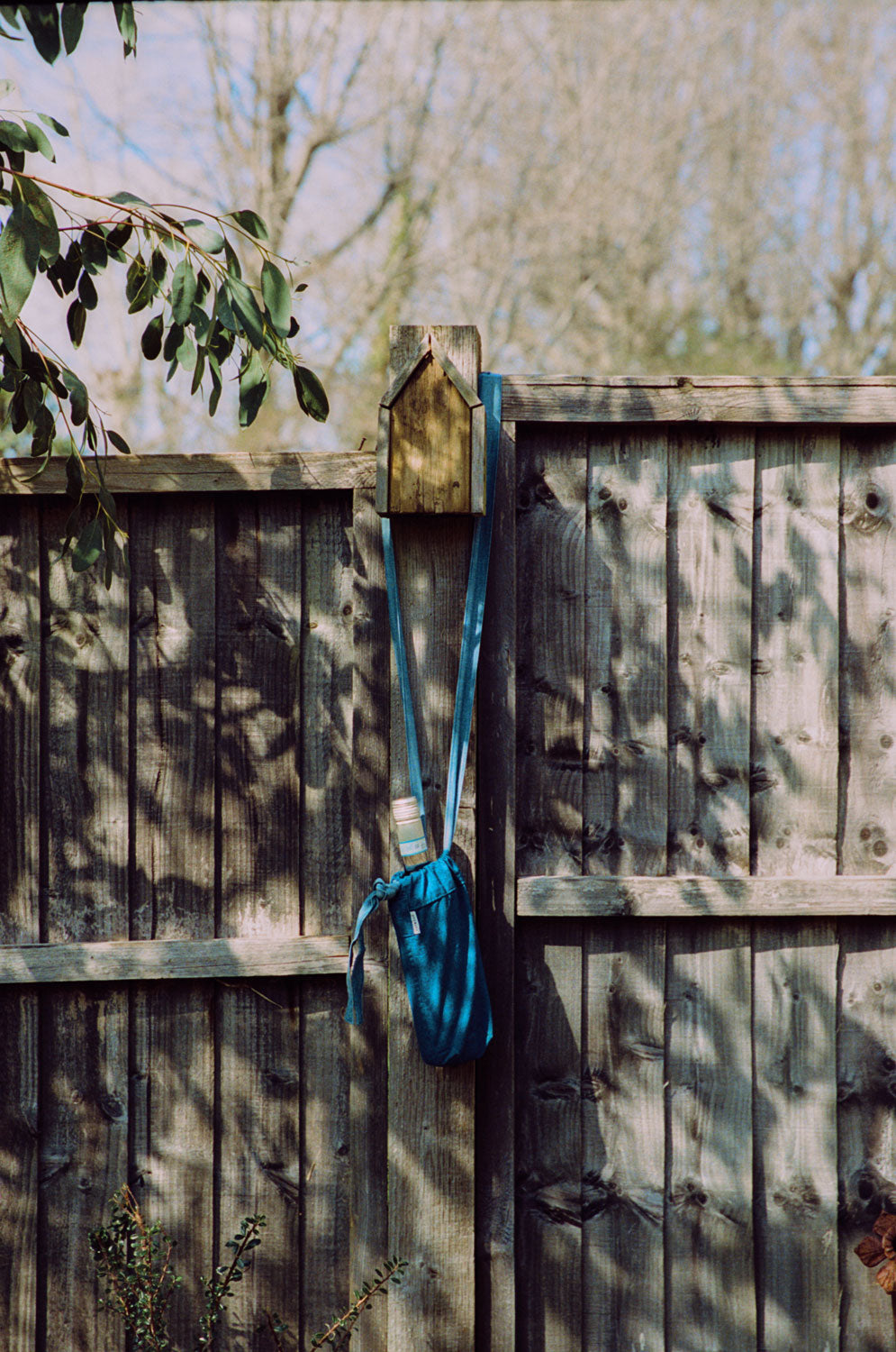 Japanese Denim Mini Bag / Bottle Bag, with a wine bottle in, hanging on a wooden fence post below a bird box. Shadows of leaves from the sunlight play on the fence.