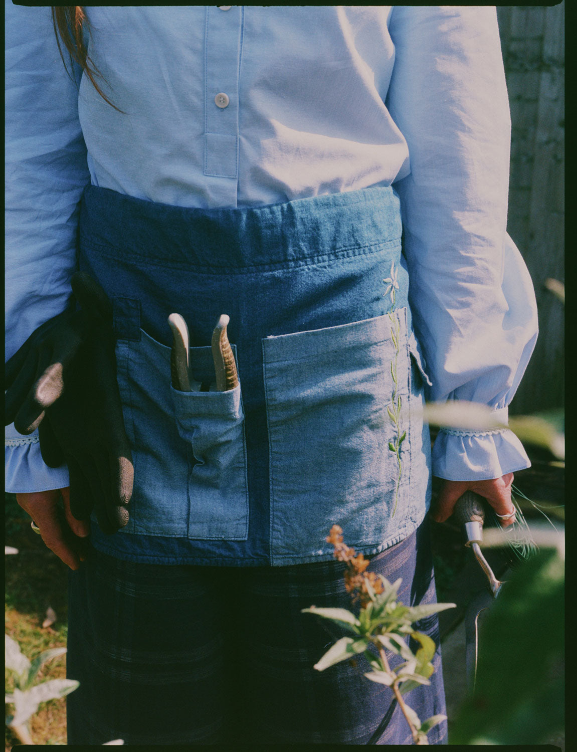 Close up of a woman standing in front of plants. She is wearings Saywood's Japanese Denim Multifunctional Gardener's Tool Belt Apron with signature flower embroidery on the pocket. The tool belt has a pair of secateurs in the pocket. The woman wears the pale blue recycled cotton Marie Blouse in top with gathered cuffs.
