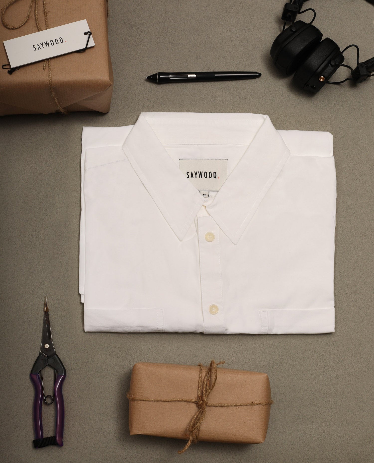 White shirt with 'Sawywood' label, brown package, scissors, pen, and headphones on a gray surface.
