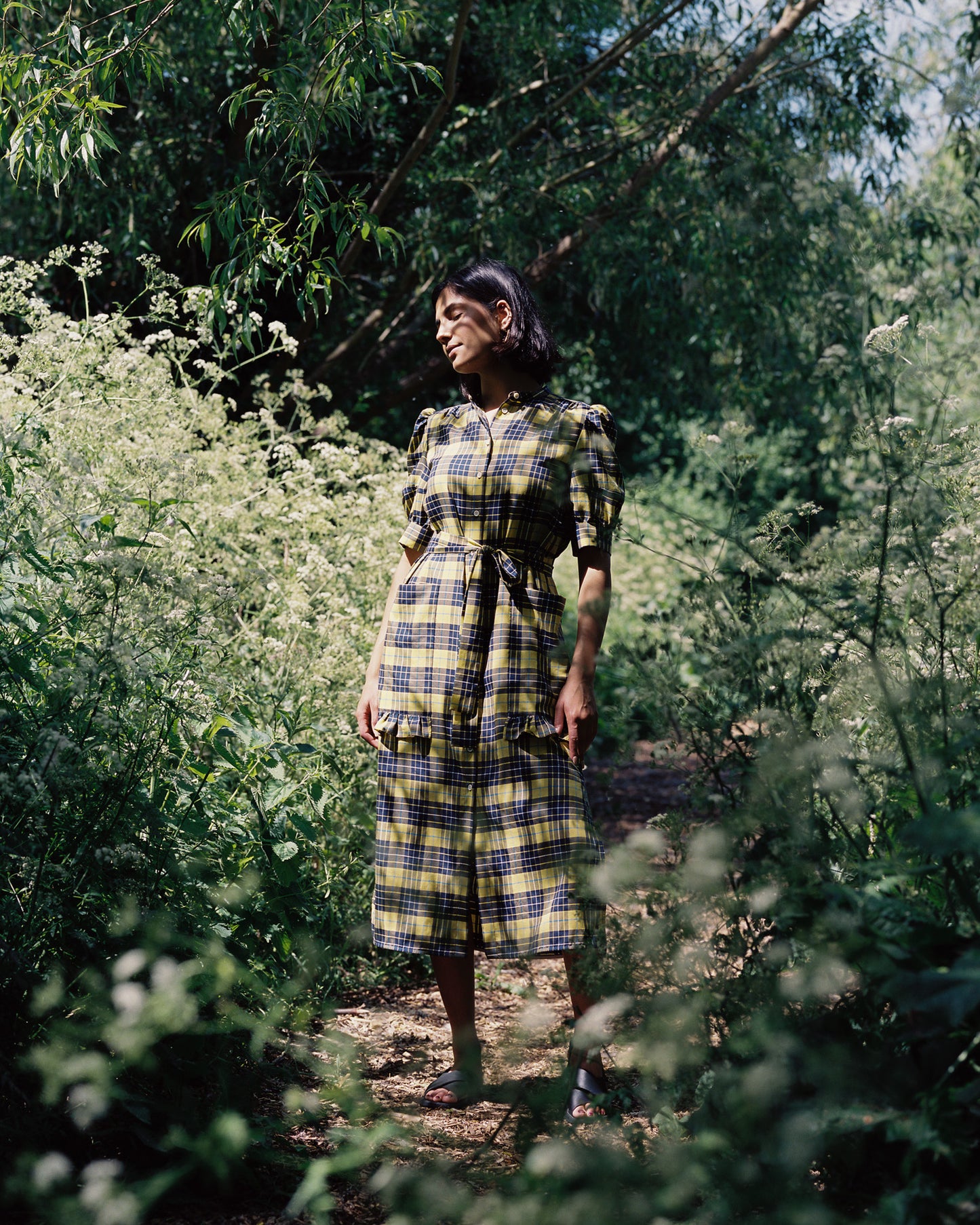 A woman stands amongst greenery and elderflower bushes, in dappled sunlight. She is wearing Saywood's Rosa Puff Sleeve Shirtdress in yellow and navy check tartan, worn with sandals. The tie belt is loosely tied around her waist.