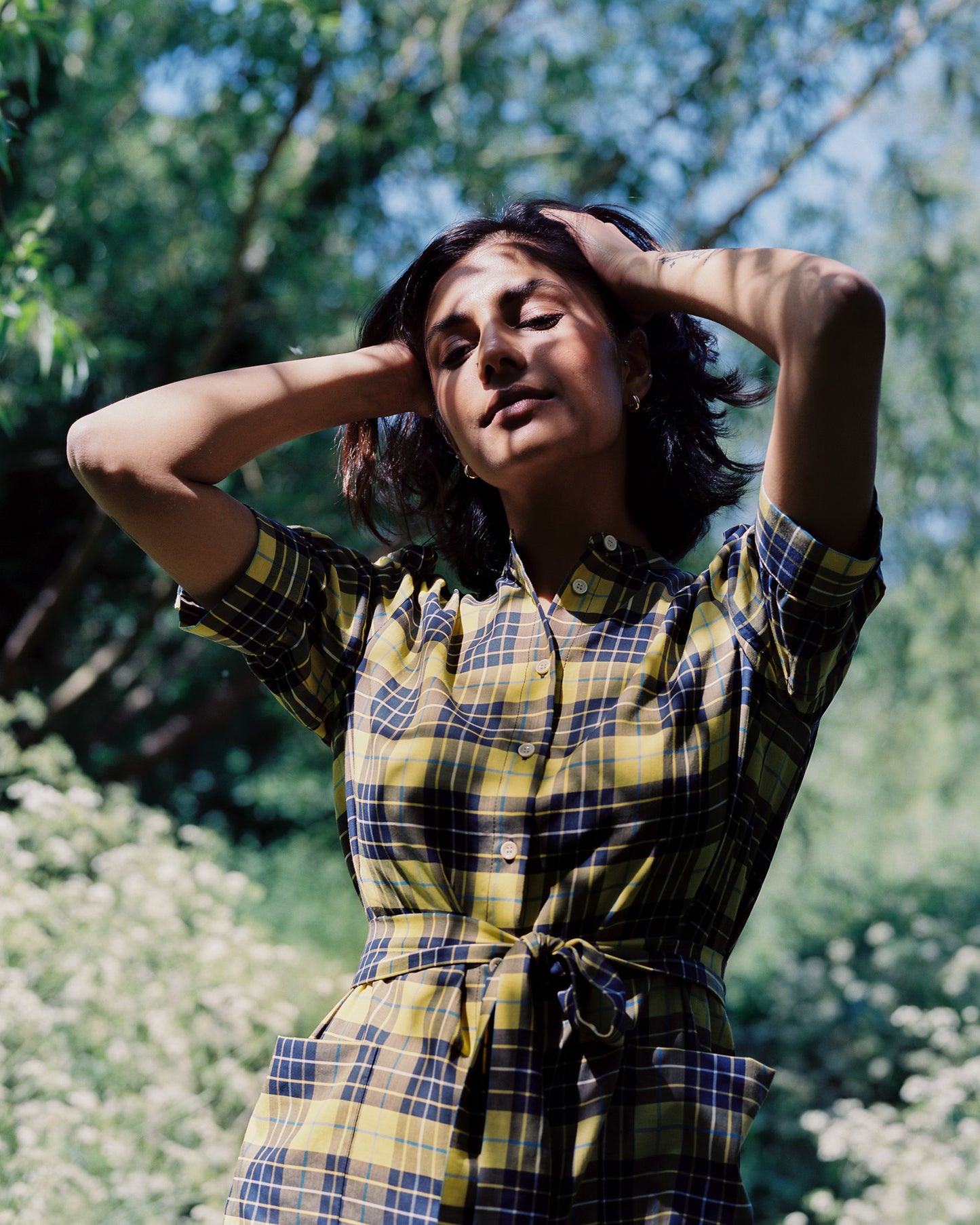 Close up of a woman standing amongst greenery and elderflower bushes, in dappled sunlight. She is wearing Saywood's Rosa Puff Sleeve Shirtdress in yellow and navy check tartan, worn with sandals. The tie belt is tied around her waist. Her hands are running through her hair on top of her head.