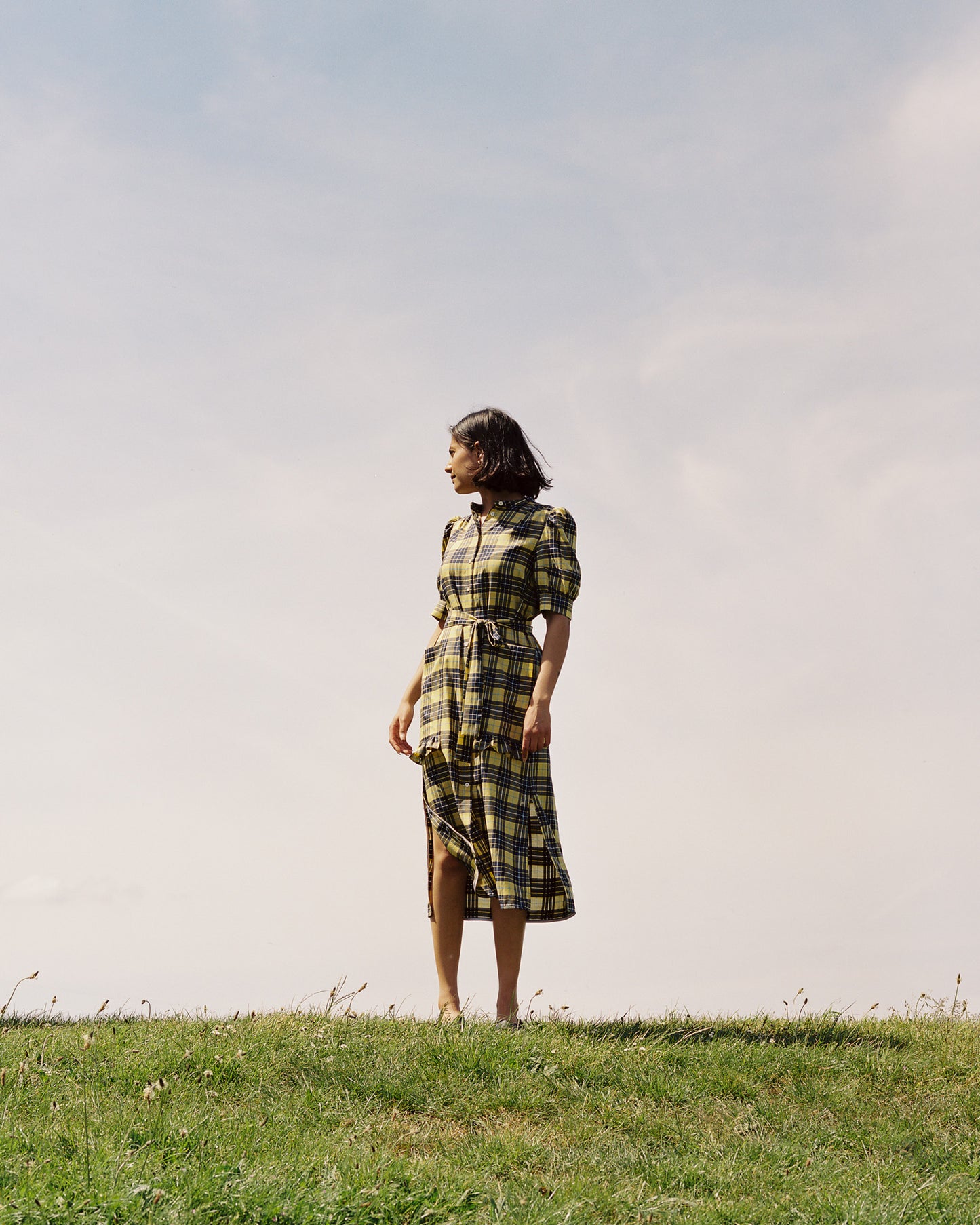 A woman stands on top of a grassy hilltop with blue sky and light whispy clouds behind her, looking back over her shoulder. She is wearing Saywood's Rosa Puff Sleeve Shirtdress in yellow and navy check tartan, worn with sandals. The tie belt is loosely tied around her waist.