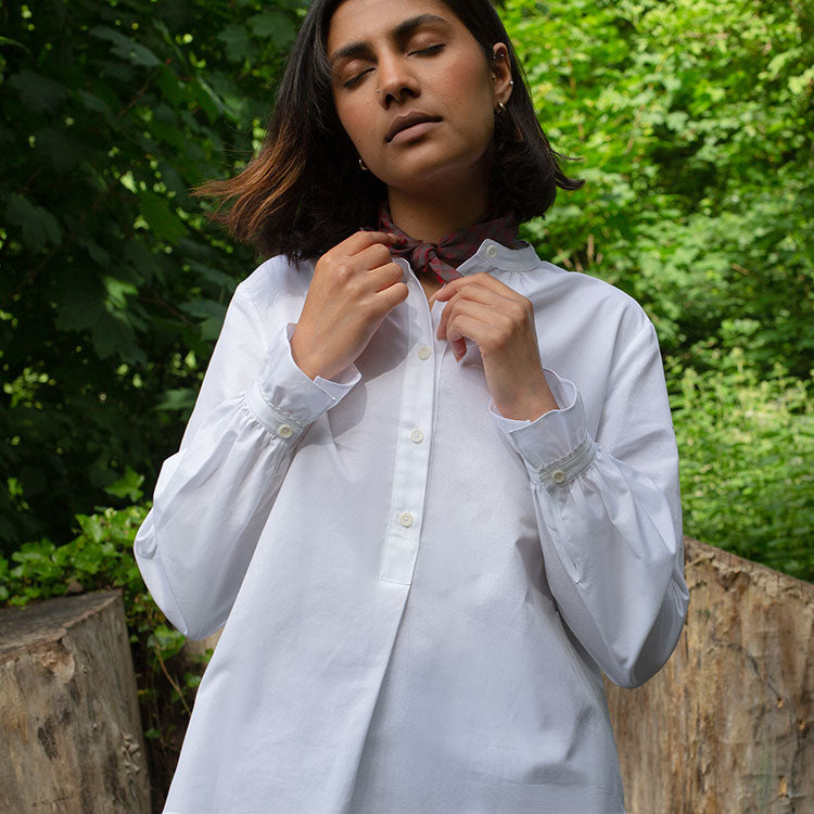 Female model is holding onto a neckerchief tied round her neck. She wears Saywood's Marie white recycled cotton shirt with lace trim. She is surrounded by trees and greenery.