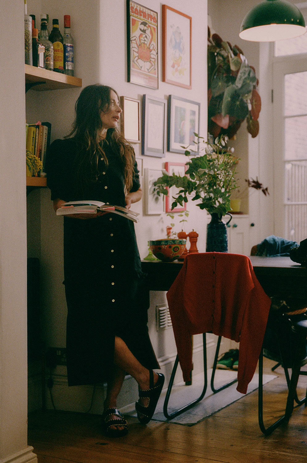 A woman stands in her kitchen diner holding an open recipe book looking out, leaning against a shelf of recipe books and drinks bottles, next to a French farmhouse style wooden table. She wears Saywood's Rosa Long Line Puff Sleeve Shirtdress in black Japanese denim, with black chunky sandals and with her hair worn down. One leg is crossed in front of the other. There are flowers on the table, pictures on the all and a red cardigan is draped over the back of a chair.