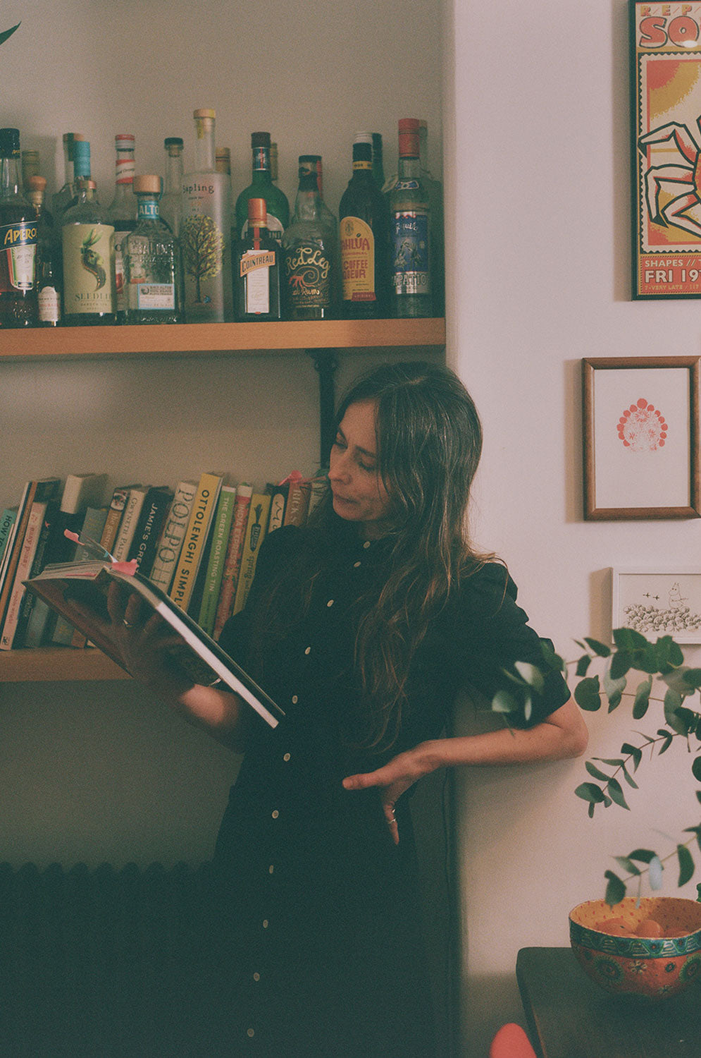 A woman with long wavy hair stands by shelves in a kitchen, reading a cook book. She is wearing Saywood's black Japanese Denim Rosa Puff Sleeve Shirtdress. She has one hand on her hip. Pictures can be seen on the wall with eucalyptus on the table at the edge of the image. the photo is shot on film.