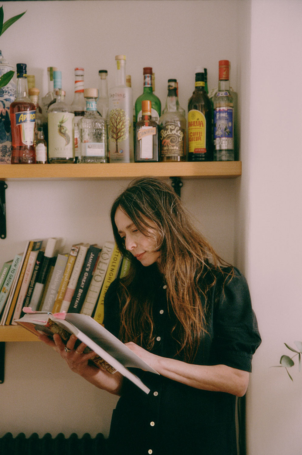 A woman with long wavy hair stands by shelves in a kitchen, reading a cook book. She is wearing Saywood's black Japanese Denim Rosa Puff Sleeve Shirtdress. The image is a close up showing from the waist up.