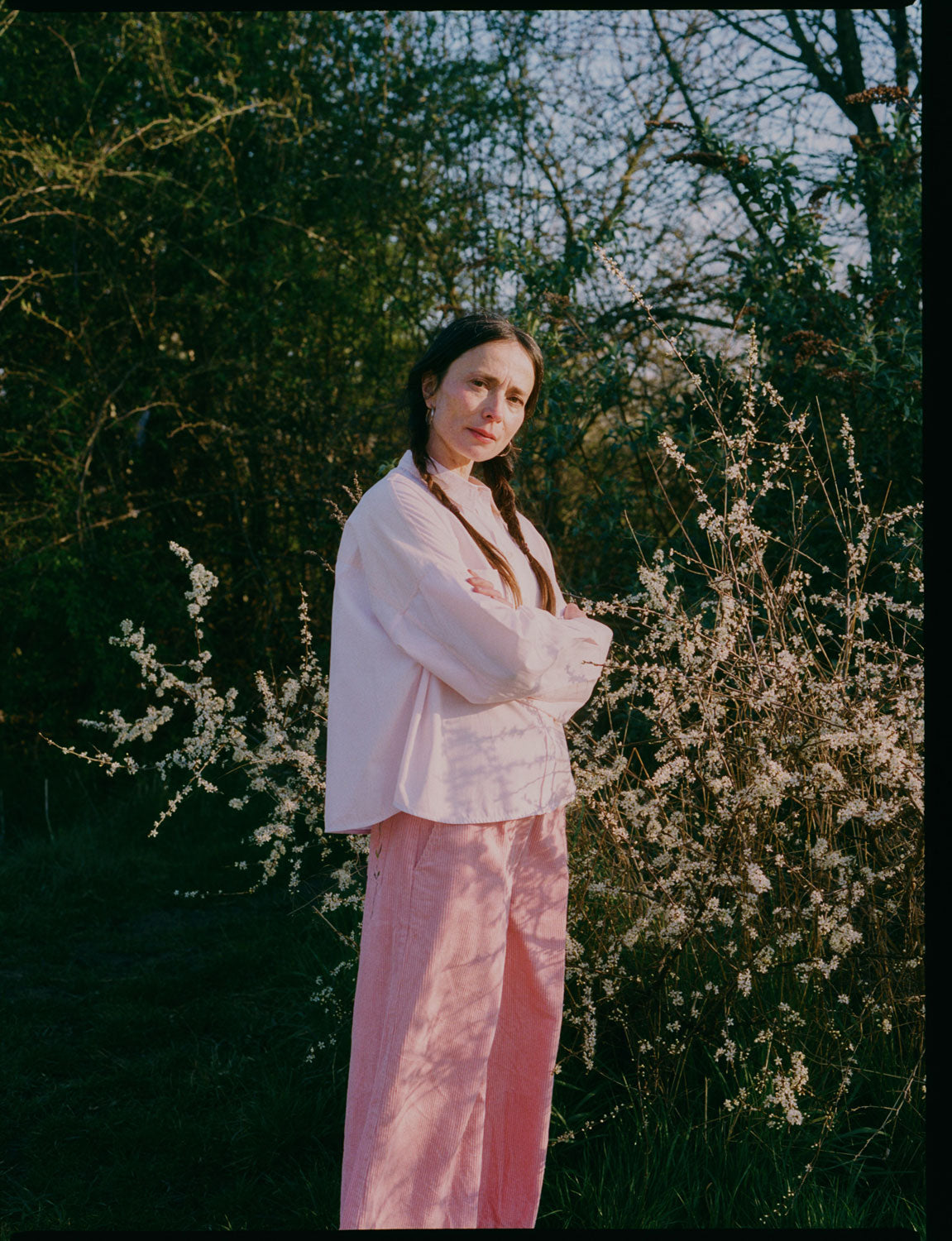 A woman stands in front of white flowering brambles, standing sideways to the camera, with her arms folded looking towards us. She wears Saywood's Amelia Wide Leg Trousers in pink corduroy with the Lela Colourblock Shirt in pink and orange. Her hair is in plaited pigtails. Image is shot on film and shows almost full length of the model.