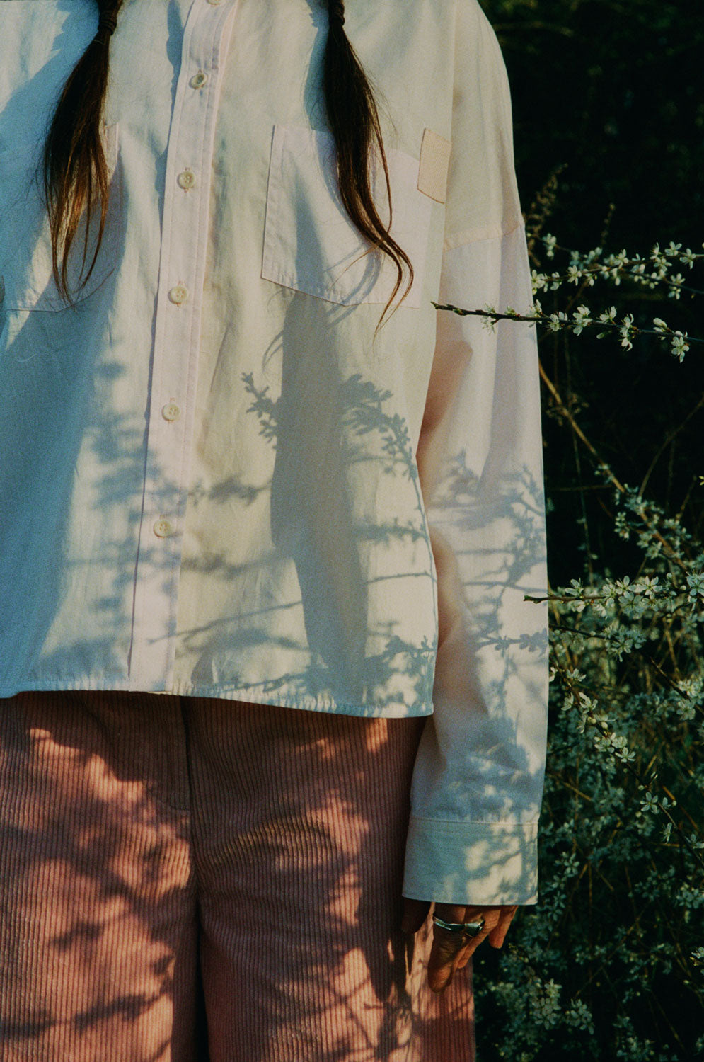 Close up of a woman standing amongst the Spring brambles, image shot on film. She wears Saywood's Lela Boxy Colourblock Shirt in pastel pink and orange, paired with the Amelia Wide Leg Trousers in pink corduroy, all made in London. Her hair is in pigtail plaits.