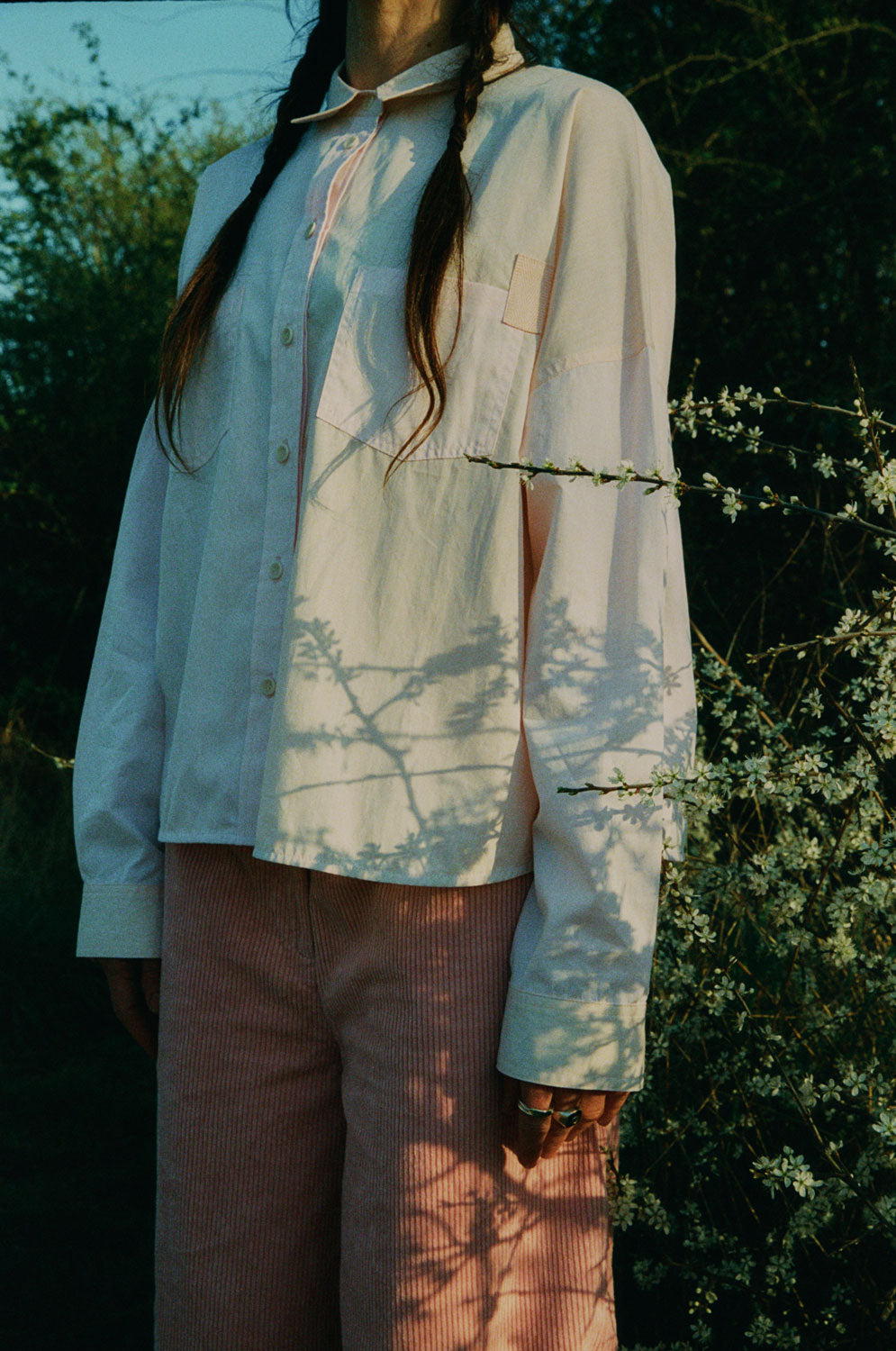 Close up of a woman standing in front of white flowered brambles. She wears Saywood's Amelia Wide Leg Trousers in pink corduroy with the Lela Colourblock Shirt in pink and orange. Her hair is in plaited pigtails. The patch pocket and herringbone tab detail can be seen on the shirt. The SAYWOOD logo is in white across the centre of the image.