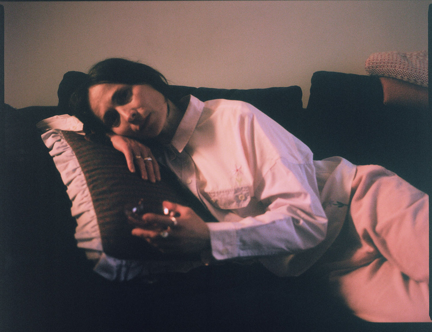 A woman is lounging on a navy sofa, leaning her head on a Saywood red check and lilac ruffle cushion. She is wearing Saywood's lilac and pink colourblock Jules Utility Shirt with the pink corduroy Amelia Trousers. The embroidered flower is visible on the check of the shirt above the pocket, and at the hem. She is holding a pink glass of wine with chunky silver rings on her fingers. Image is shot on film.