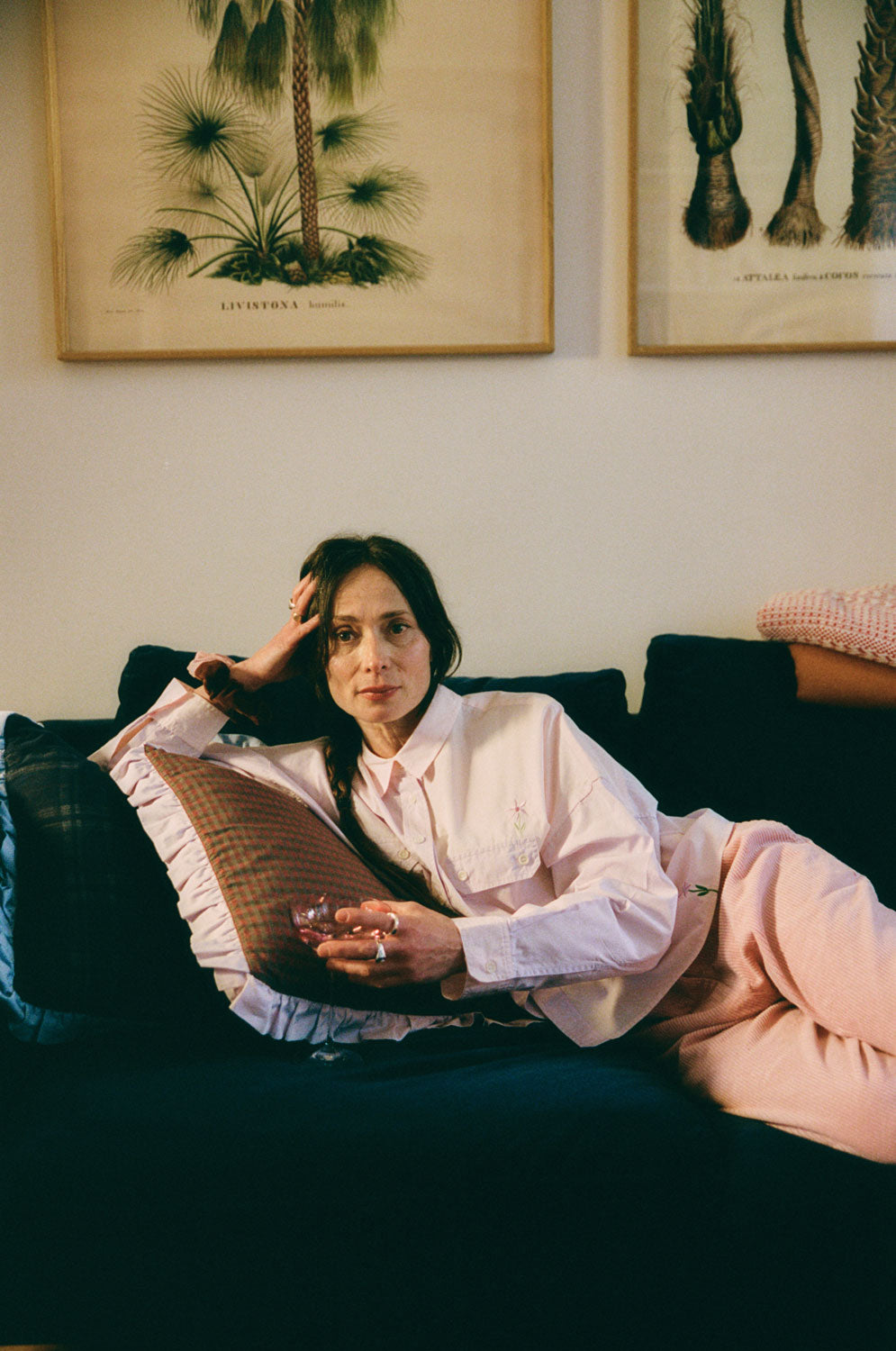 A woman is lounging on a navy sofa, leaning on a Saywood red check and lilac ruffle cushion. She is wearing Saywood's lilac and pink colourblock Jules Utility Shirt with the pink corduroy Amelia Trousers. The embroidered flower is visible on the check of the shirt above the pocket, and at the hem. She is holding a pink glass of wine with chunky silver rings on her fingers. Pictures of retro illustrated palm trees are hanging on the wall behind her.