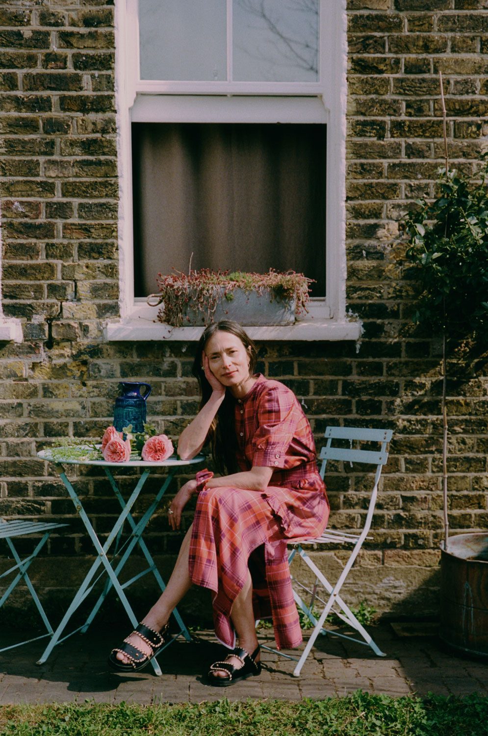 A woman sits on a blue garden chair by a table with flowers on in front of a window in the garden. She is wearing Saywood's Rosa Puff Sleeve Shirtdress in coral, purple and red check with black sandals, with her head resting on her hand.