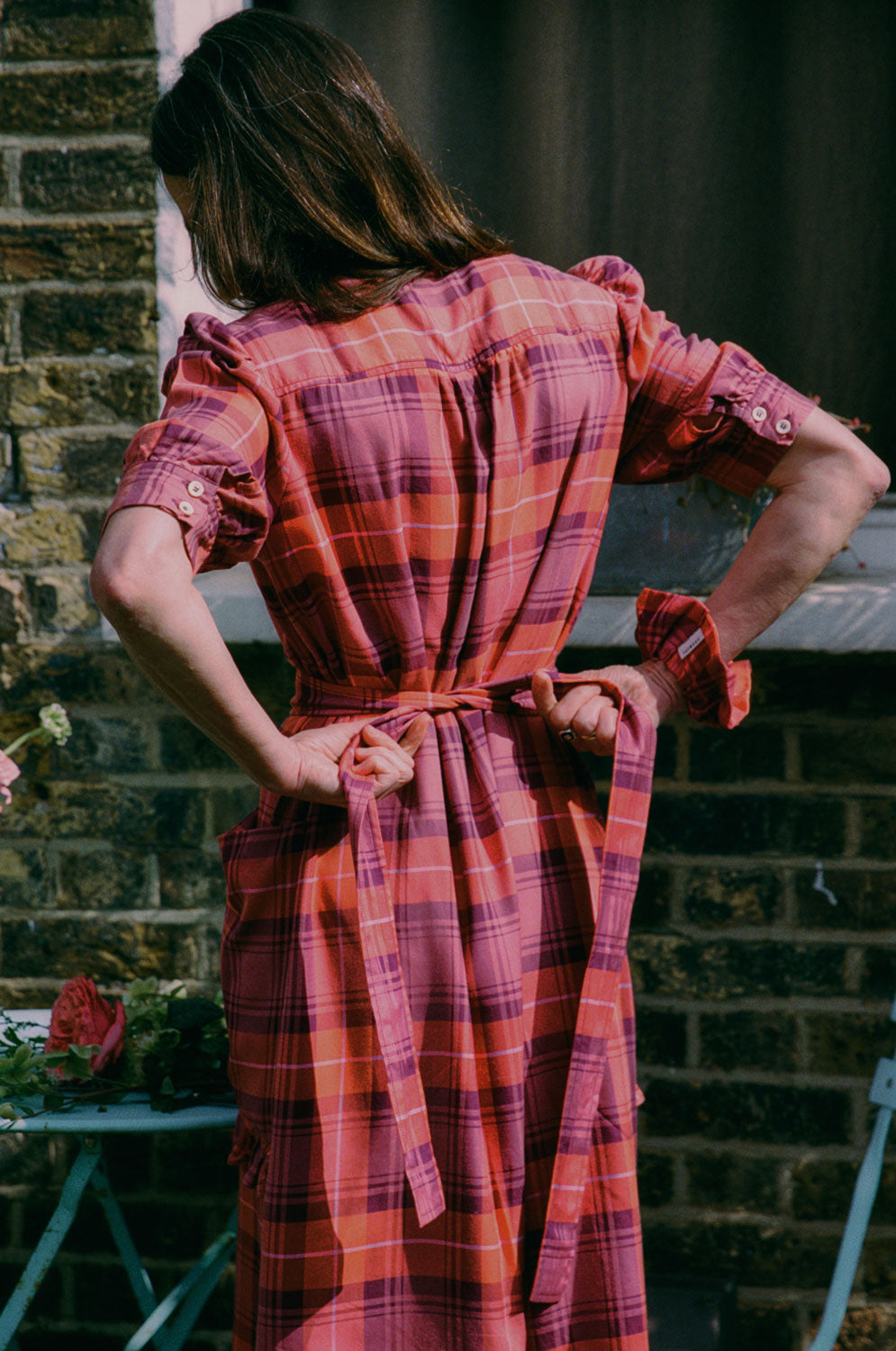 A woman with long hair stands with her back to the camera, tying a bow in the tie belt at the back of her dress. She is wearing Saywood's Rosa Puff Sleeve Shirtdress in coral, red and purple check, with a matching scrunchie on her wrist. Made in London, UK. Flowers can be seen on the table.