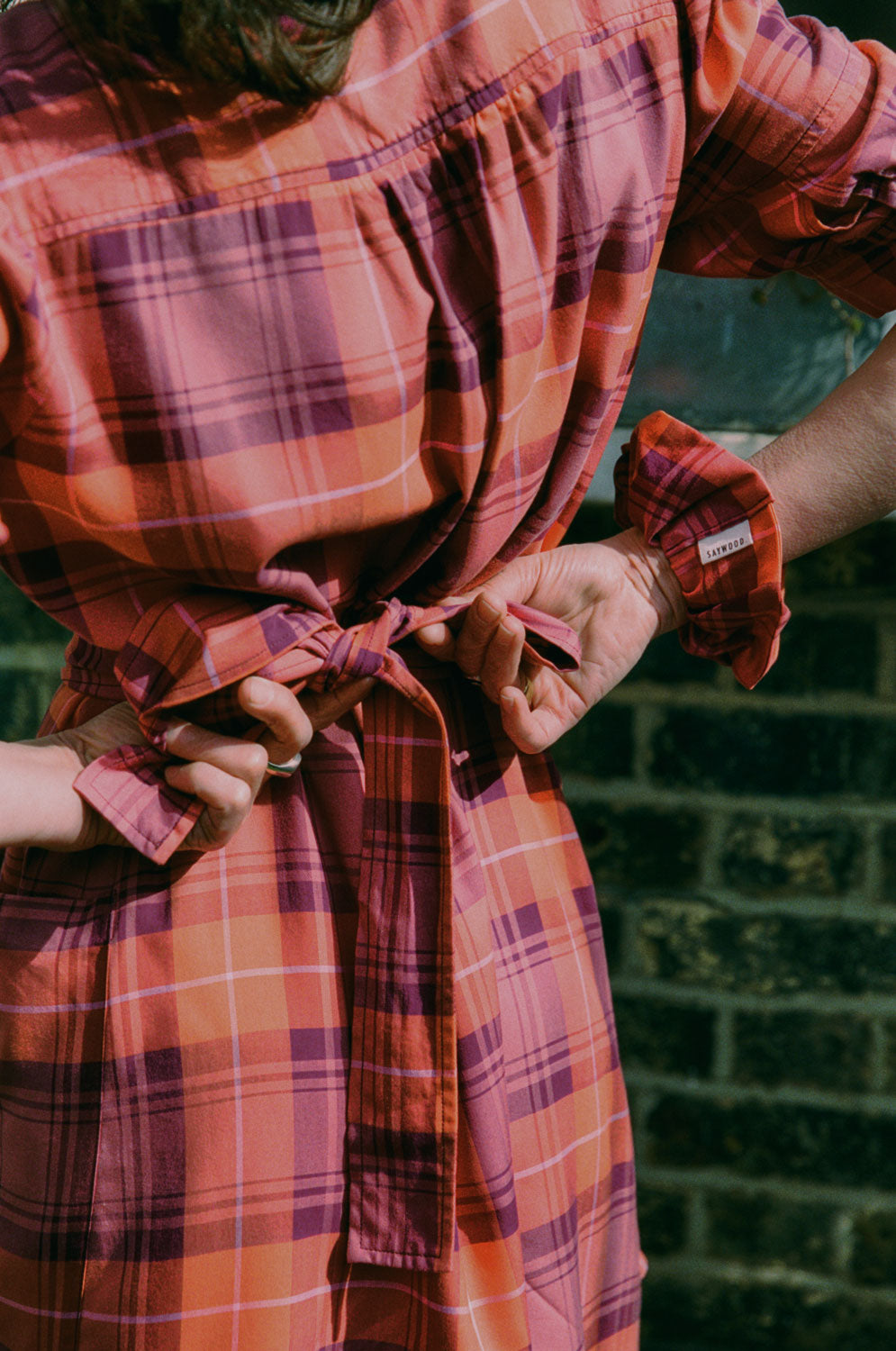 Close up. A woman with long hair stands with her back to the camera, tying a bow in the tie belt at the back of her dress. She is wearing Saywood's Rosa Puff Sleeve Shirtdress in coral, red and purple check, with a matching scrunchie on her wrist. Made in London, UK.