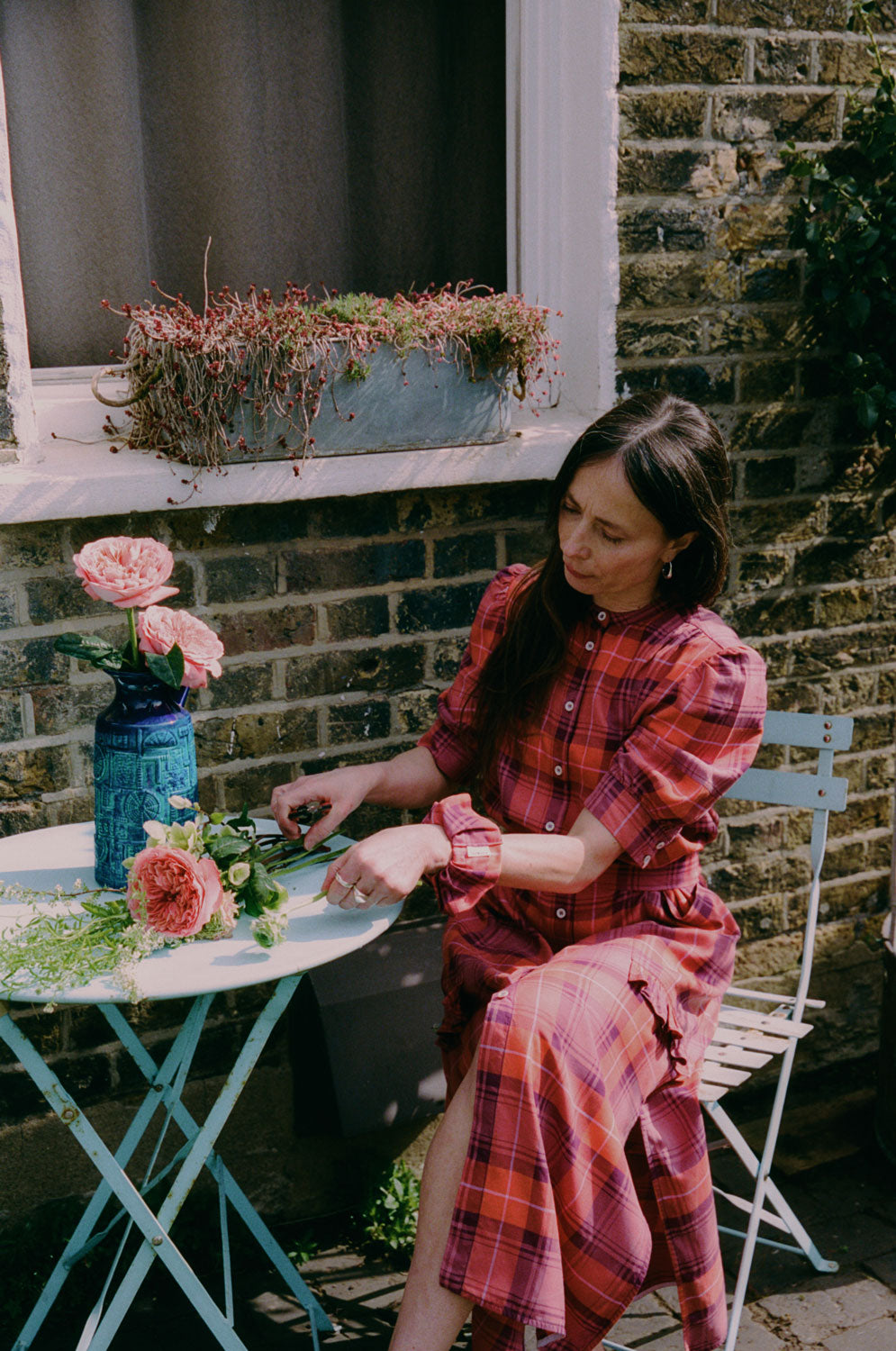 A woman sits at a blue garden table preparing pink flowers in a blue vintage vase. She is wearing Saywood's Rosa Puff Sleeve Shirtdress in coral red check with a matching scrunchie on her wrist.