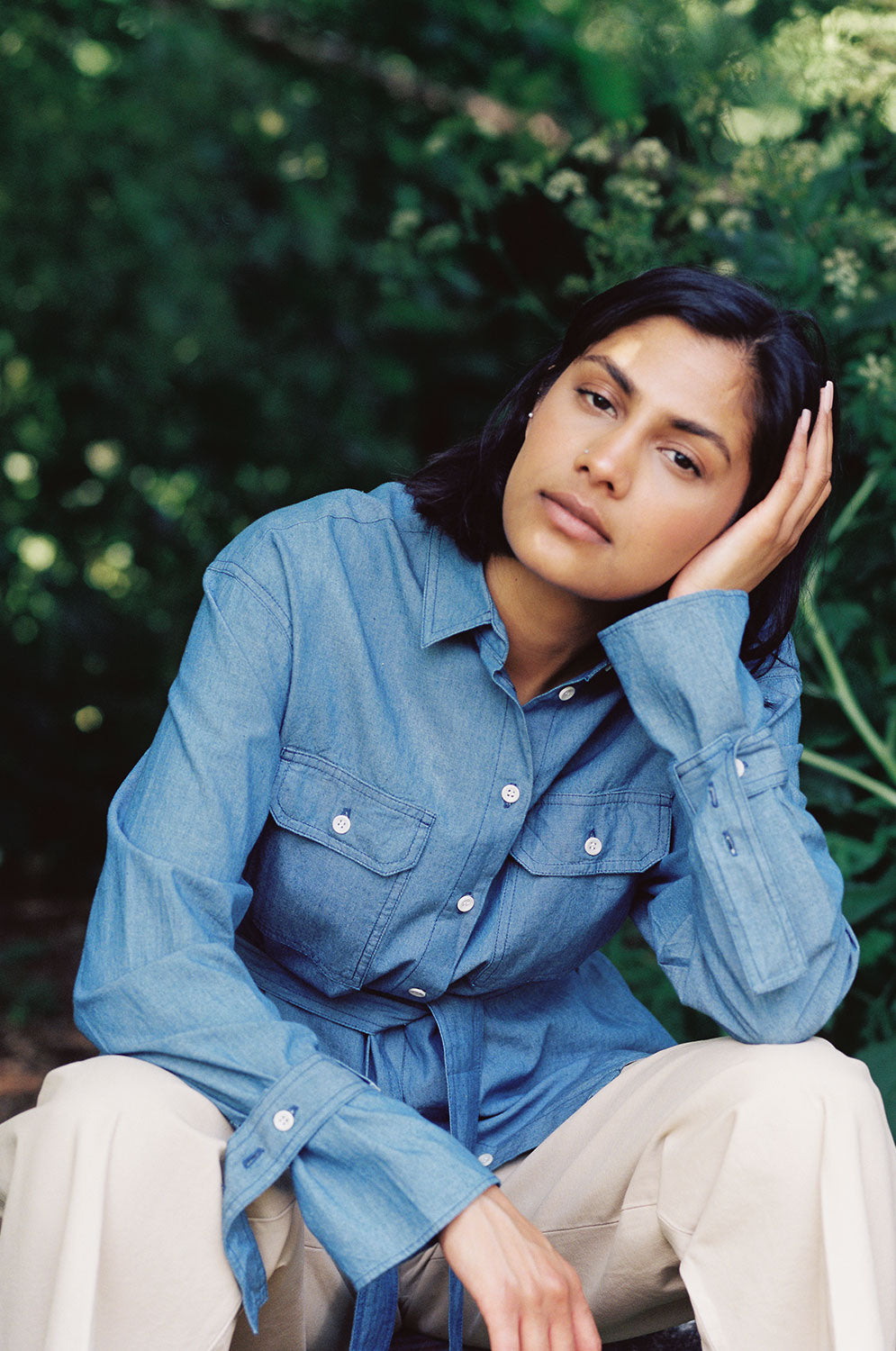 Close up of a woman sitting on a log amongst leafy trees, with her head resting on one hand. She is wearing Saywood's Zadie Oversized Shirt in lightwash Japanese denim, with the tied belt tied around the waist. The utility patch pockets, top stitch detailing and cuff belts can be seen.