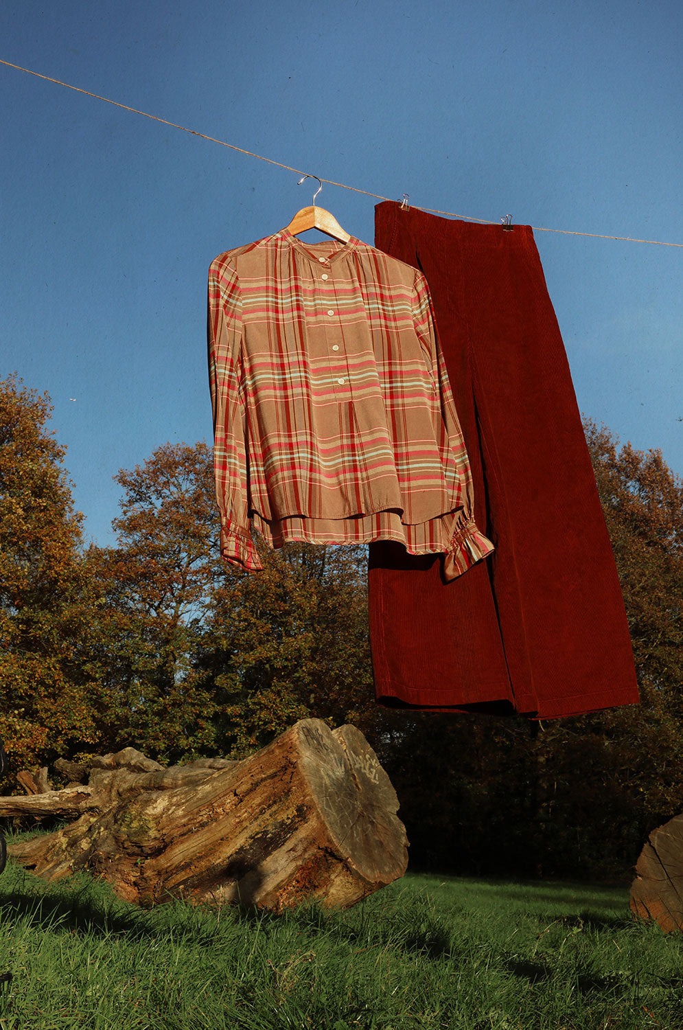 Hanging on a line in the middle of a field surrounded by trees, with a big fallen tree trunk is the background, are Saywood's Amelia Wide Leg Trousers, full length leg, in caramel tan organic cotton corduroy, and the Marie pink and hazelnut check Blouse.