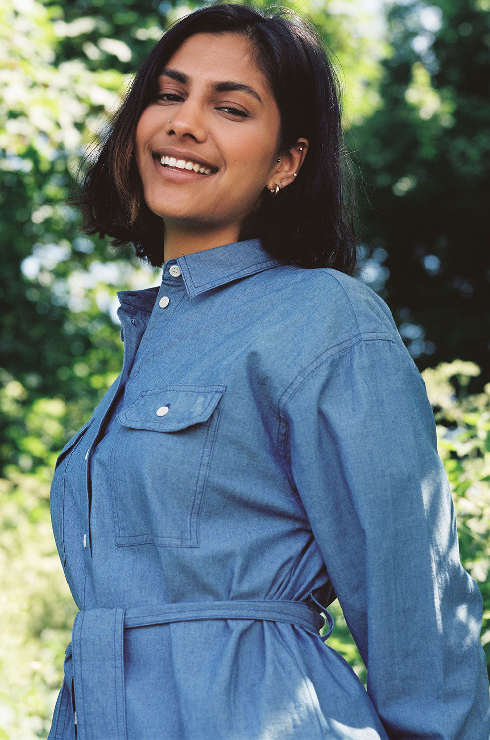 Close up of a woman standing amongst leafy trees, turned at a slight angle. She is wearing Saywood's Zadie Oversized Shirt in lightwash Japanese denim, with the tied belt tied around the waist. The utility patch pockets and top stitch detailing can be seen.