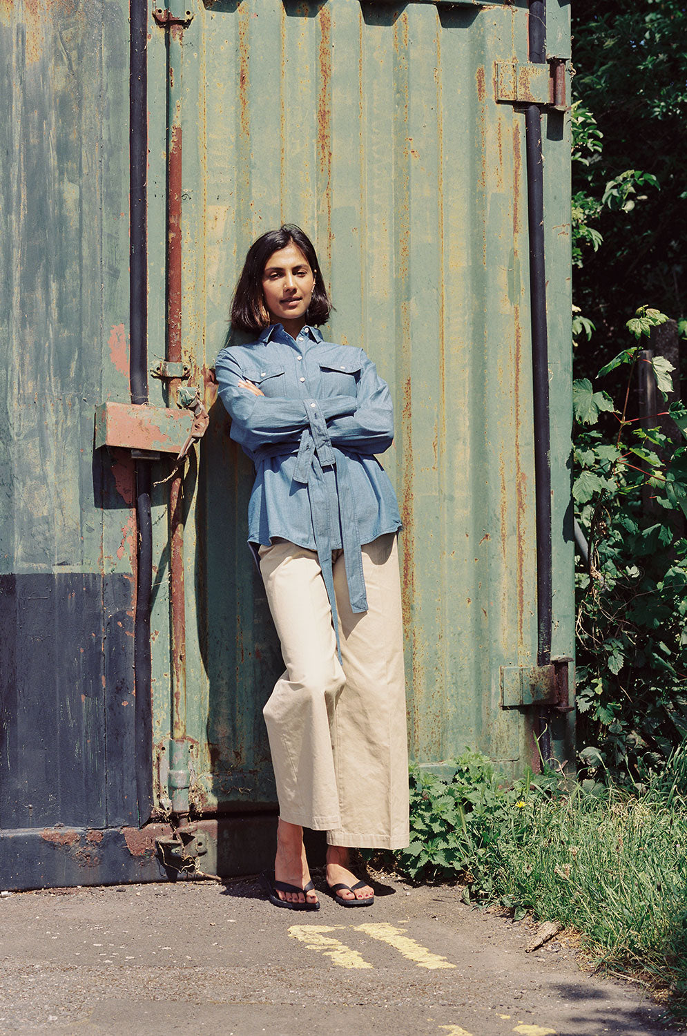 Model leans against a green shipping container, with arms crossed in front of her waist. She wears Saywood's Zadie Boyfriend Shirt in Japanese Denim with a pair of beige trousers and black sandals.