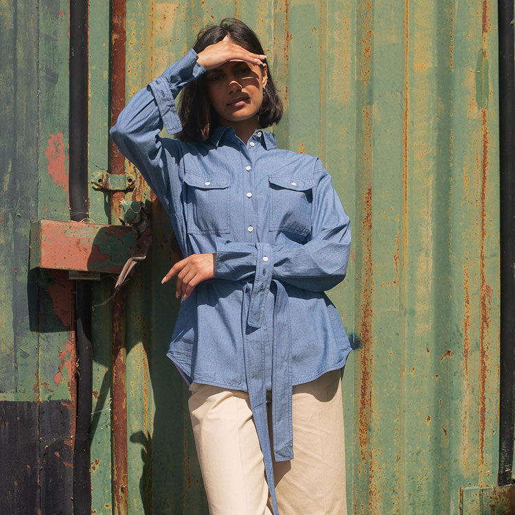 Model leans against a green shipping container, with one hand shielding her eyes from the sun. She wears Saywood's Zadie Boyfriend Fit Shirt with safari styling in Japanese denim, and taupe trousers.