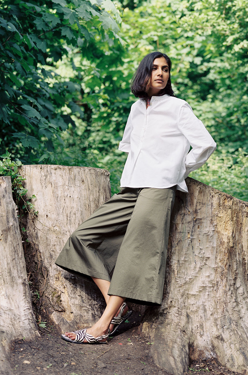Model leans against a tree stump, one knee bent with hands behind her back on the tree, with greenery in the background. She wears Saywood's Amelia wide leg trouser in khaki cotton sateen, with the white recycled cotton Marie A-line Blouse with a red check neckerchief tied round her neck. Worn with black and white zebra print sandals.