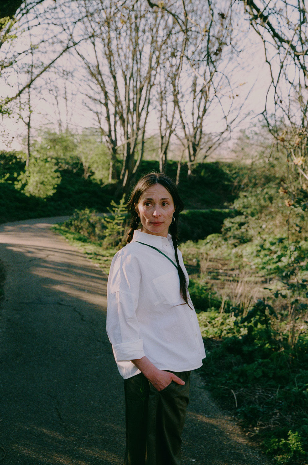 A woman is standing on a path in a green park surrounded by nature. She is turned to the side looking over her shoulder with her hand in her trouser pocket. She wears Saywood's Amelia Wide Leg Culotte Trousers in khaki, with the white recycled cotton Lela Boxy Shirt. Her hair is plaited in pigtails and she has a green cross body bag across her.