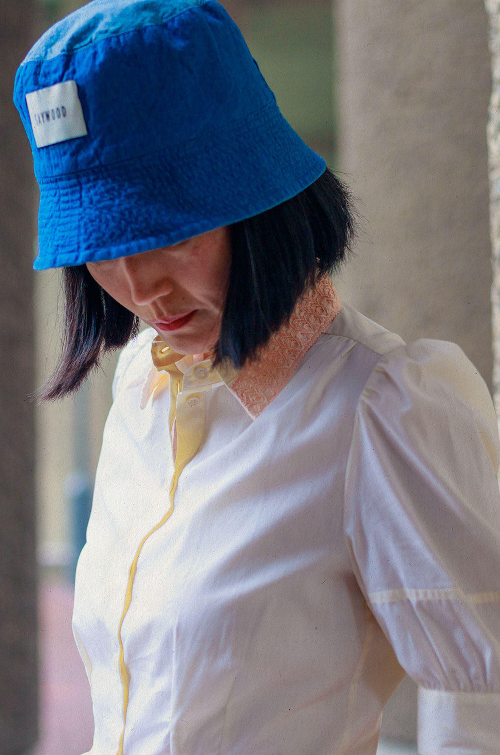 Close up of a woman with dark hair, looking downwards. She is wearing Saywood's Joni Puff Sleeve Blouse in lemon, with fit and flare shaping, peach lace collar and scalloped hem. Worn with the Japanese denim Bucket Hat.