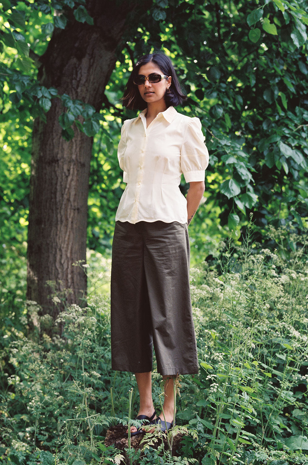 A woman stands on a tree stump, with her hands behind her back, surrounded by nature. She is wearing Saywood's Joni Puff Sleeve Blouse, with fit and flare darting, scallop hem adn lace trim on the collar. Worn with the Amelia Wide Leg Trousers in khaki cotton, sunglasses and black sandals.