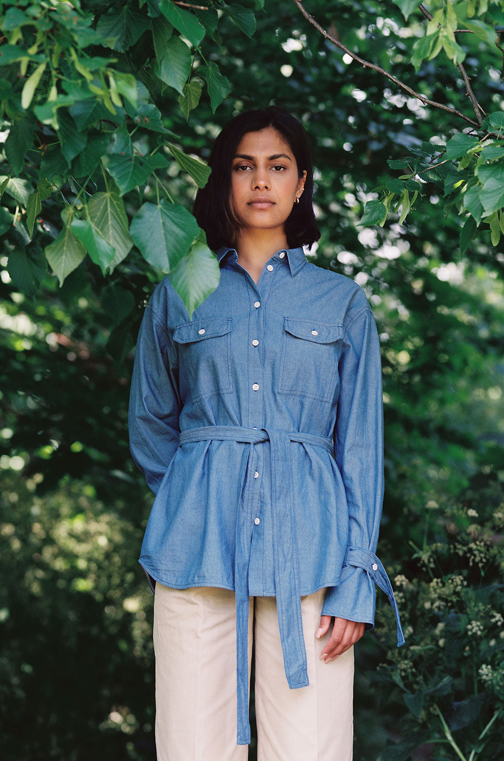 A woman stands amongst leafy trees, with one arm behind her back. She is wearing Saywood's Zadie Boyfriend Fit shirt in lightwash Japanese denim, worn with light tan chinos.