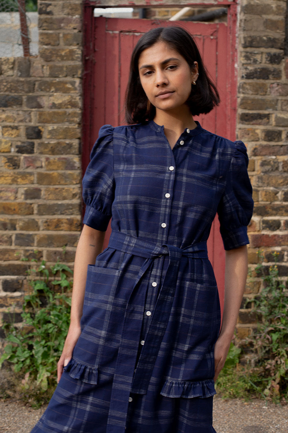 A woman stands in front of a brick wall and red wooden door. She is wearing Saywood's Rosa Puff Sleeve Shirtdress in navy check with the belt loosely tied around her waist. The gathered puff sleeves and patch pockets with ruffle trims can be seen in the close up shot.