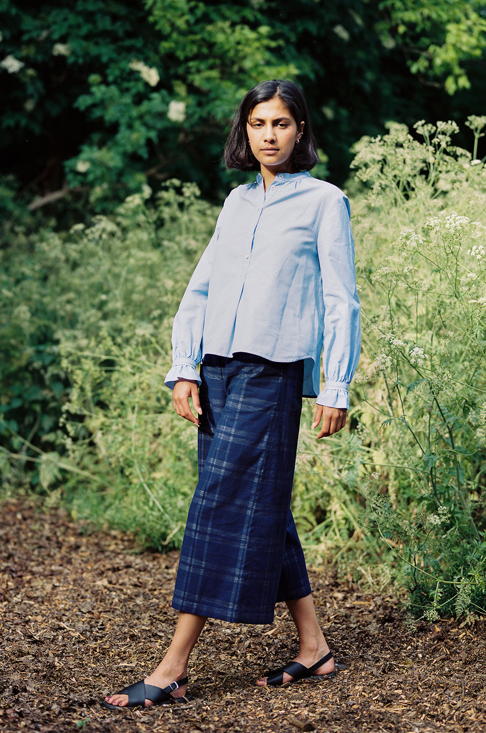Full length shot of model wearing Saywood's Amelia check trousers, wide leg culotte shape in navy check, with black sandals. Worn with the pale blue shirt, the Marie a-line blouse. She is standing outside surrounded by wild plants and greenery.