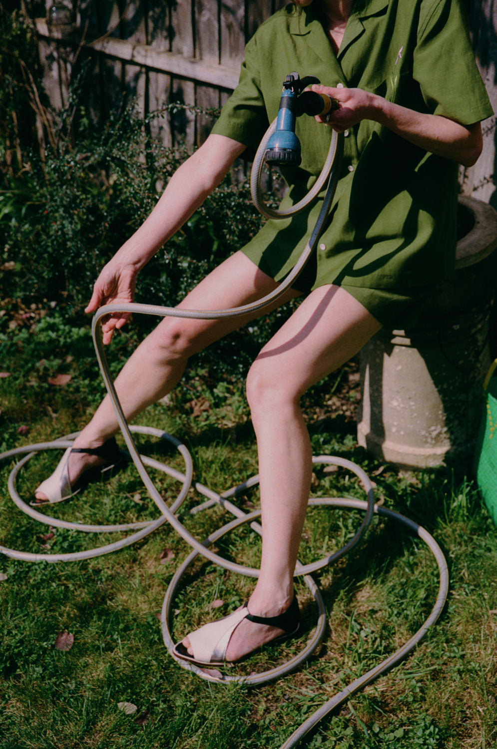 A woman is perched on a garden pot, holding a garden hose, which unravelling on the grassy ground, with a wooden fence behind her. She is wearing the Frances Unisex Cuban Shirt in olive green, with short sleeves and the signature embroidered flower in lilac on the check pocket. Worn with the Bessie A-line Shorts in olive green as a matching twin set. The image is shot slightly from above.