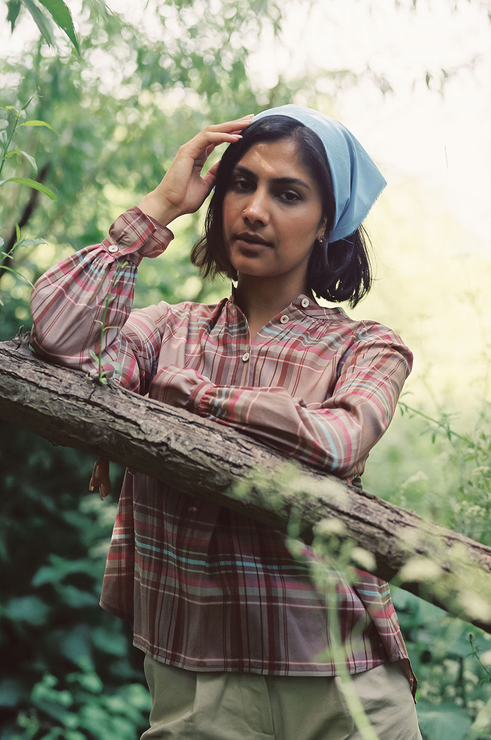 A woman is leaning on a tree branch, at an angle across the image, with greenery in the background. She is wearing Saywood's Marie A-line Gathered Neck Blouse in pink and hazelnut check, worn with tan chinos and a recycled cotton pale blue bandana in her hair. One arm rests on the tree trunk whilst her other hand is touching her head. The gathered frill cuffs can be seen.