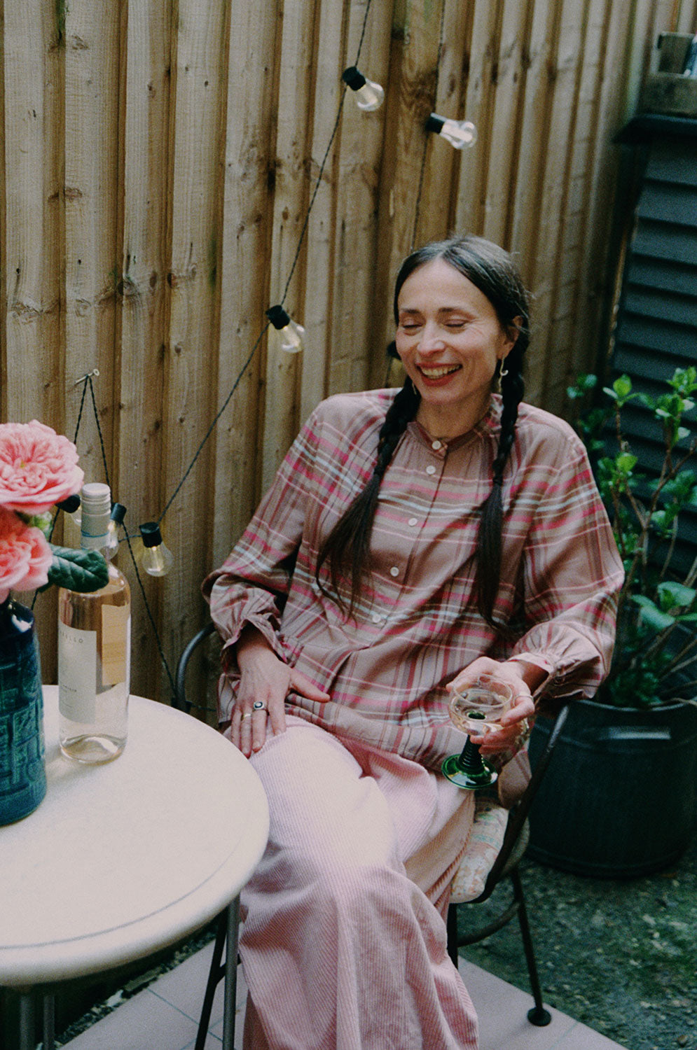 A woman is sitting in a iron garden chair holding a glass of wine, with a small round marble table in front of her, with a bottle of wine on it and a blue vase with large pink rose flowers. She is wearing Saywood's pink and hazelnut check Marie A-line Blouse with gathered neck details and gathered frill cuffs, with the Amelia Wide Leg Culotte Trousers in pink cotton corduroy. She's smiling, with two plaited pigtails in her hair. Behind her is a wooden fence with outdoor lights strung to it and a plant.