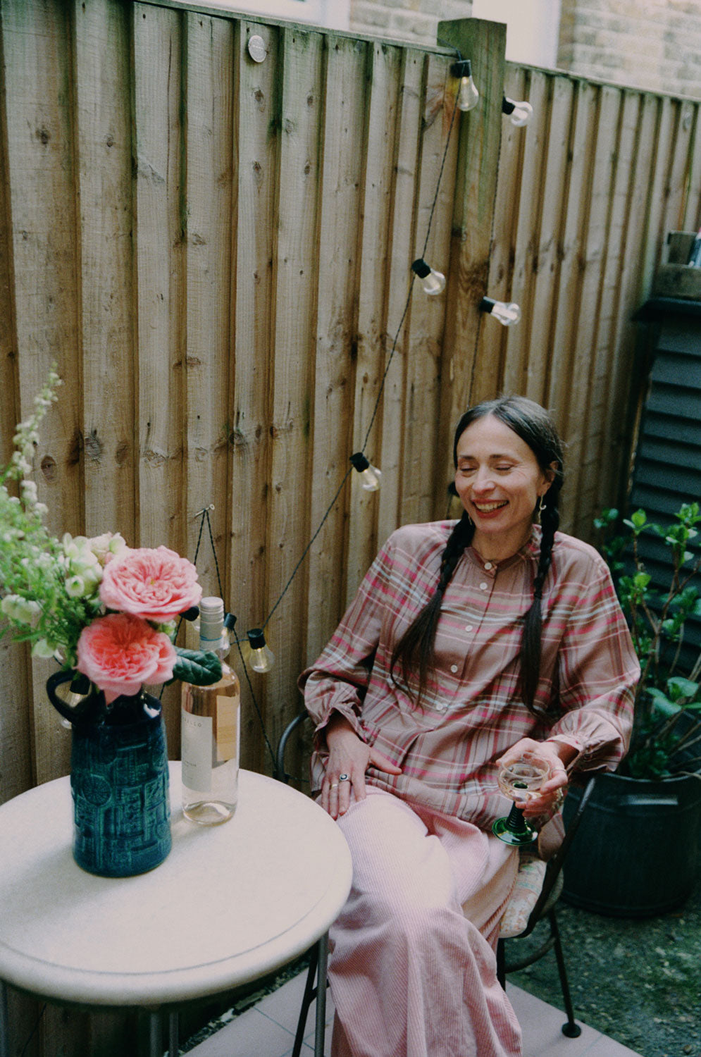 A woman is sitting in a iron garden chair holding a glass of wine, with a small round marble table in front of her, with a bottle of wine on it and a blue vase with large pink rose flowers. She is wearing Saywood's pink and hazelnut check Marie A-line Blouse with gathered neck details and gathered frill cuffs, with the Amelia Wide Leg Culotte Trousers in pink cotton corduroy. She's smiling, with two plaited pigtails in her hair. Behind her is a wooden fence with outdoor lights strung to it and a plant.