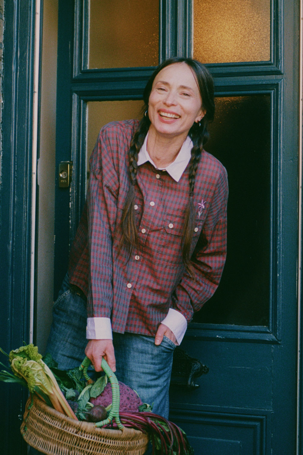 Close up of a woman standing, smiling, in front of a teal blue front door, peering round the brick wall of the porch arch. She holds a wicker basket of vegetables, with her hair in plaited pigtails. She wears Saywood's Jules Utility Shirt in red micro check with contrast lilac collar and cuffs, worn with mid wash jeans.
