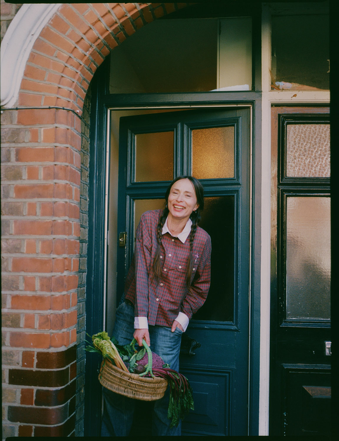 A woman stands, smiling, in front of a teal blue front door, peering round the brick wall of the porch arch. She holds a wicker basket of vegetables, with her hair in plaited pigtails. She wears Saywood's Jules Utility Shirt in red micro check with contrast lilac collar and cuffs, worn with mid wash jeans.