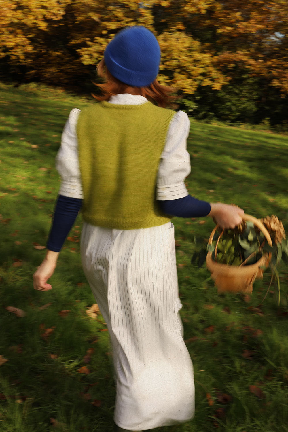 A woman is walking with her back to the camera, surrounded by trees, holding a wooden garden basket with flowers in. She is wearing Saywood's white pinstripe shirtdress, the Rosa Puff Sleeve Shirtdress, with a navy long sleeve top underneath and a green sweater vest layered over the top, with a blue beanie hat. The image is slightly blurred as she moves.