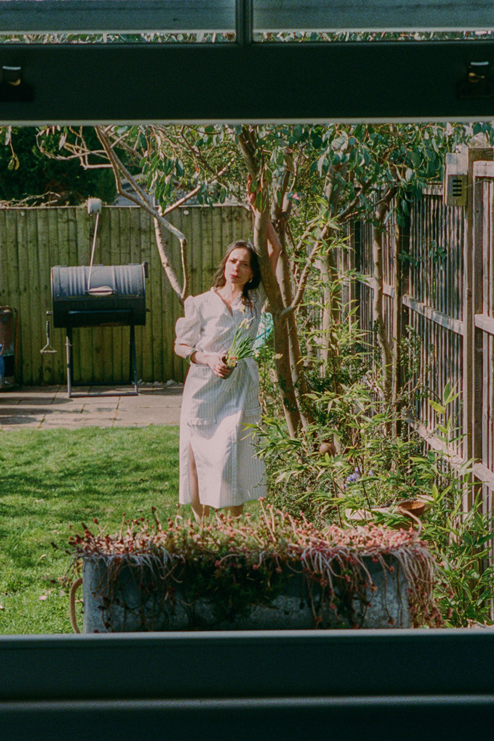 A woman stands in a garden, by a eucalyptus tree, holding a daffodil plant. She is wearing Saywood's white cotton pinstripe Rosa Puff Sleeve Shirtdress with the tie belt tied round her waist, with the patch pockets with the ruffle trim just visible. She wears her hair down, and is leaning against the eucalyptus tree with one arm up leaning on the tree. The image is shot through an open window.