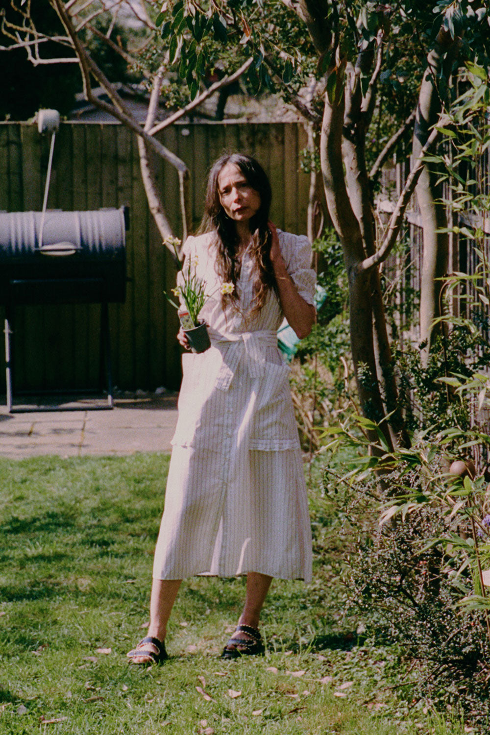 A woman stands in a garden , by a eucalyptus tree, holding a daffodil plant. She is wearing Saywood's white cotton pinstripe Rosa Puff Sleeve Shirtdress with the tie belt tied round her waist, with the patch pockets with ruffles at the bottom edge. She wears her hair down, with black sandals on her feet, and one hand touching her long hair.