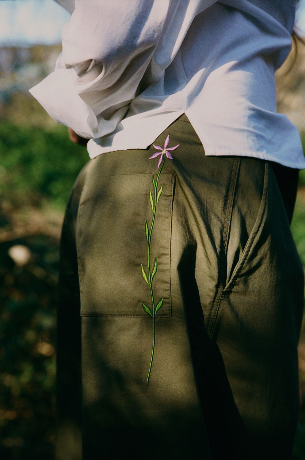 Close up detail of the model with her arms crossed behind her back, showing the side split at the hem of the white recycled cotton Lela Shirt. The signature Saywood embroidered flower in lilac on the back pocket of the khaki Amelia Wide Leg Trousers can be seen in the centre of the image.